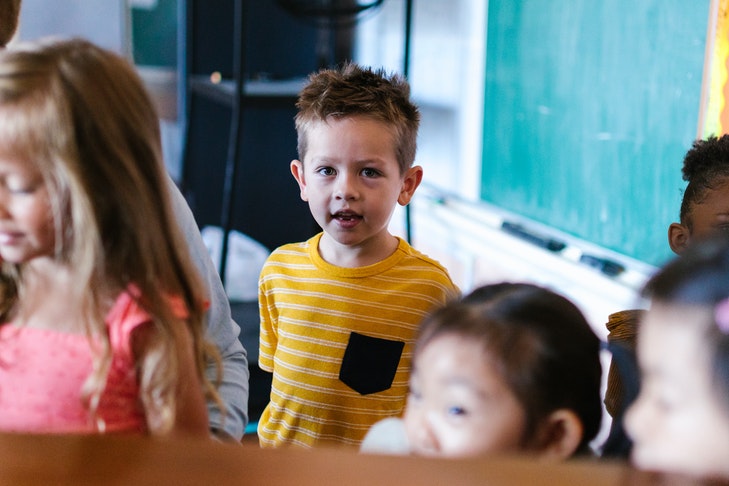 Ein Junge steht in einem Klassenzimmer vor einer Tafel, umgeben von Mitschülerinnen.