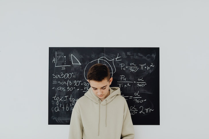 Ein Junge steht mit gesenktem Kopf vor einer Tafel, auf der Mathematikaufgaben angeschrieben sind.