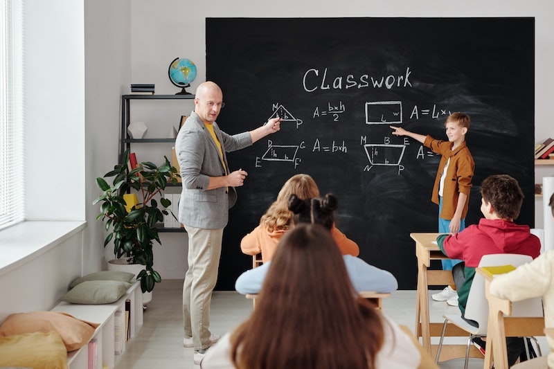 Ein Mathematiklehrer und ein Schüler stehen vor der Tafel und zeigen mit dem Finger auf geometrische Formen, die anderen SchülerInnen hören interessiert zu