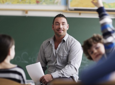 Im Vordergrund ist ein Schüler, der seine Hand hebt, im Hintergrund sitzt ein Lehrer und lacht